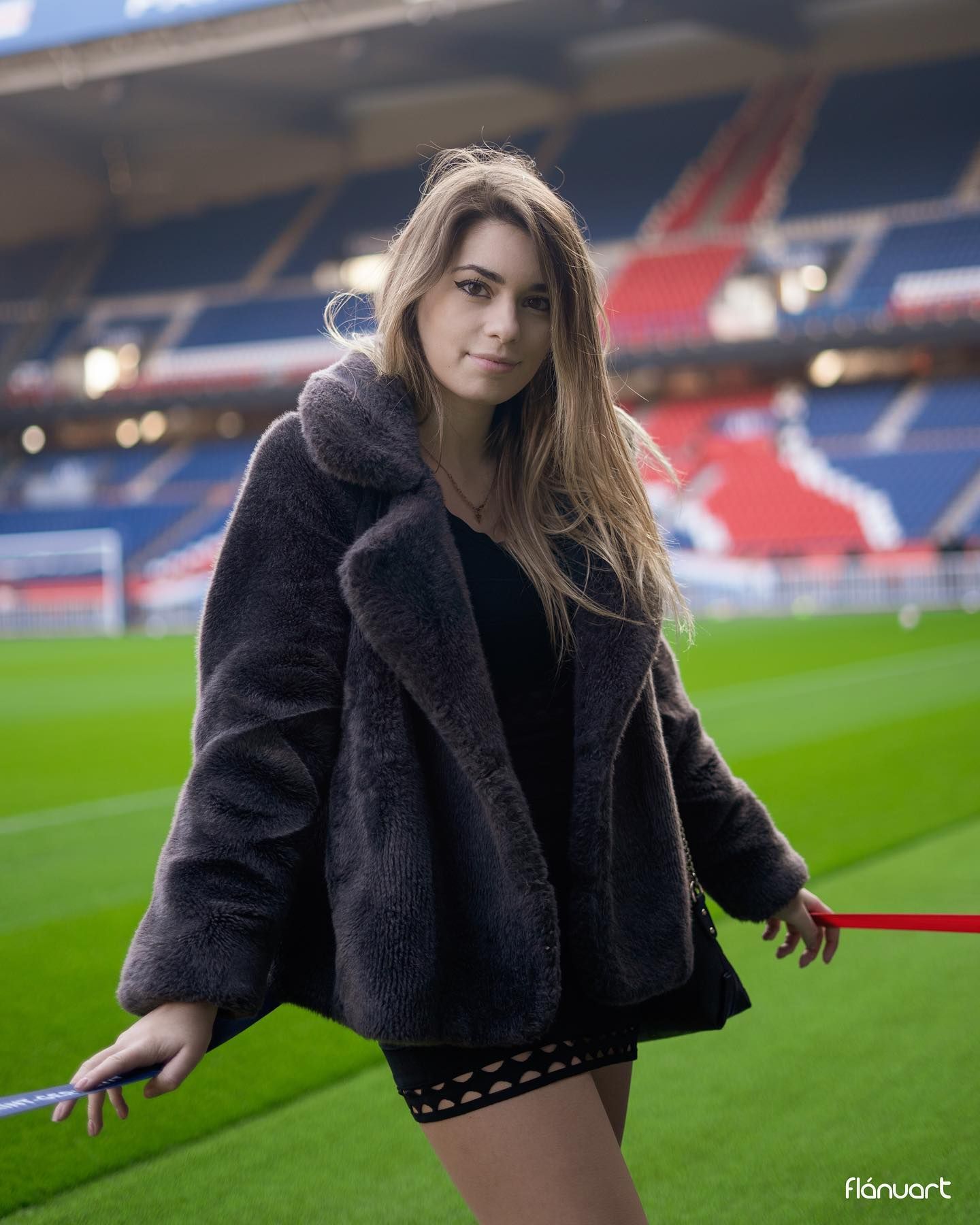 Young woman in gray teddy coat poses on a football stadium pitch holding a red barrier.