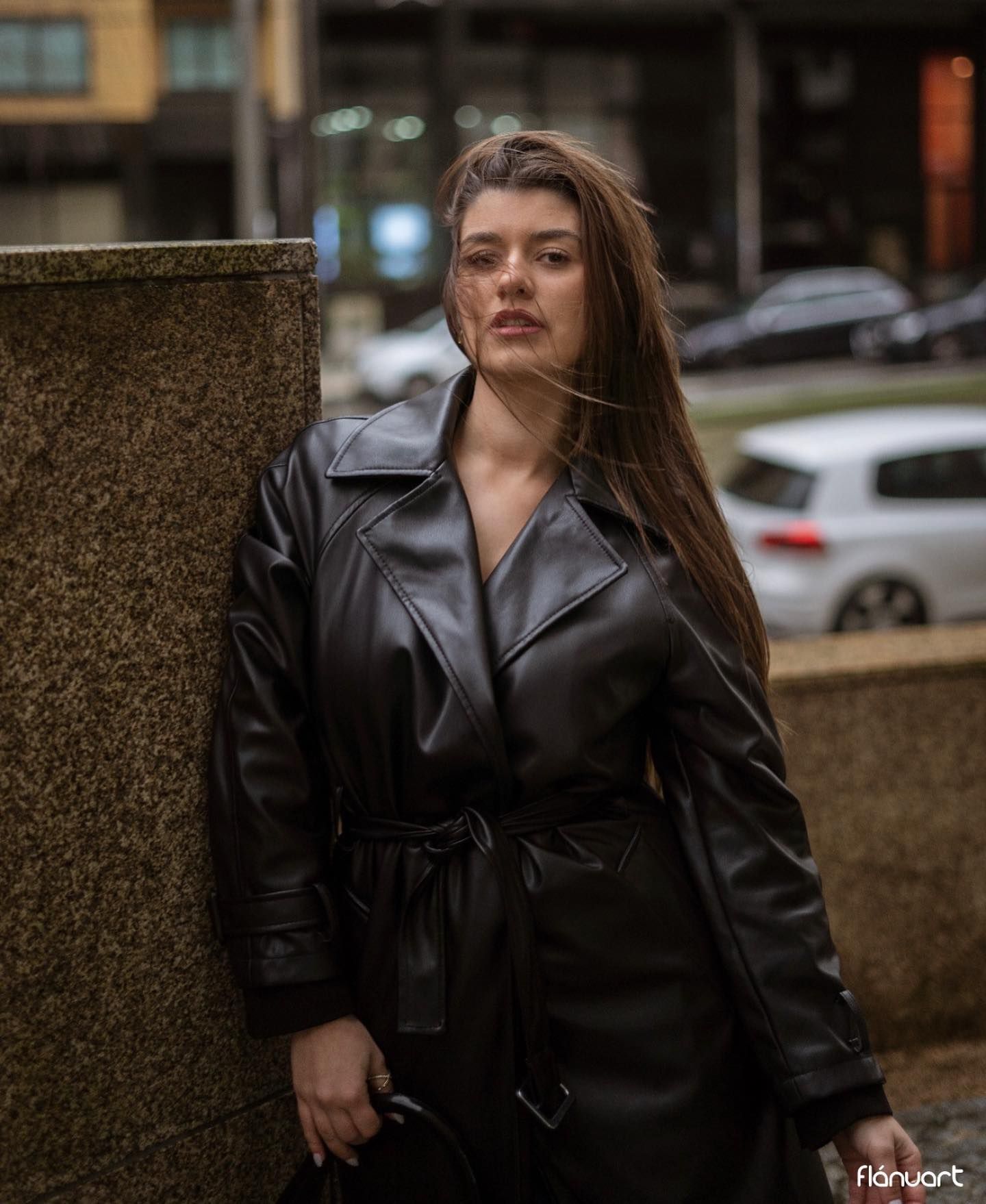 Woman in black leather trench coat leaning against urban stone column.