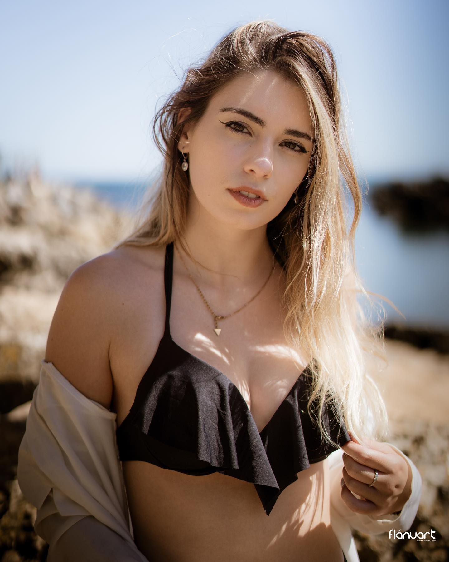 Blonde woman in black ruffle bikini top posing on rocky coastal background.