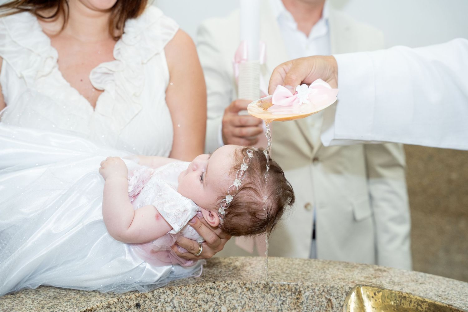 Priest pouring holy water over infant during baptism ceremony, with parents in formal white attire.