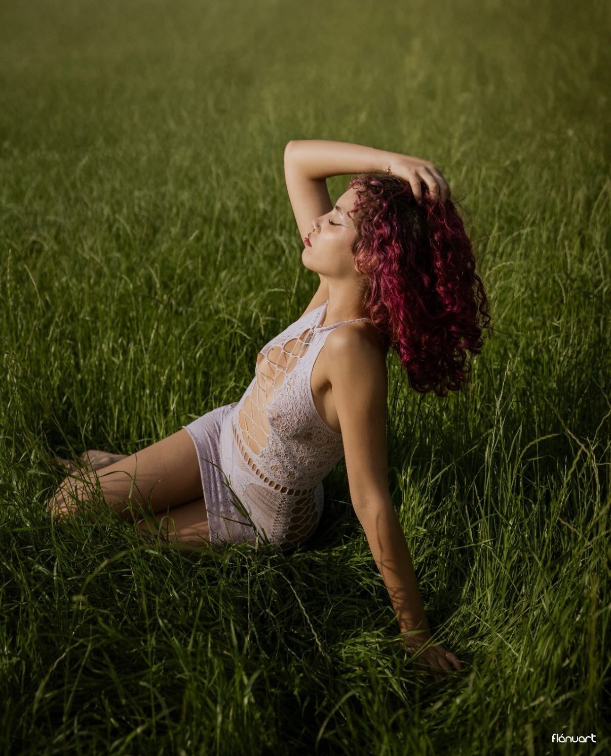 Young woman with red curly hair posing in lush green grass field wearing white lace outfit.