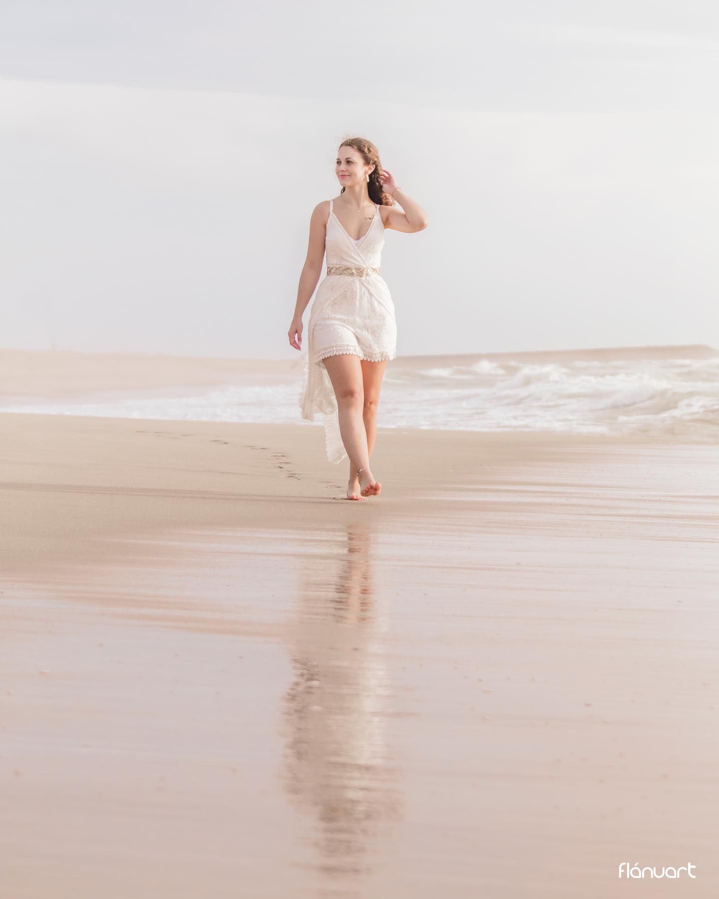 Woman in white dress walking along a calm shoreline with gentle waves on a cloudy day.