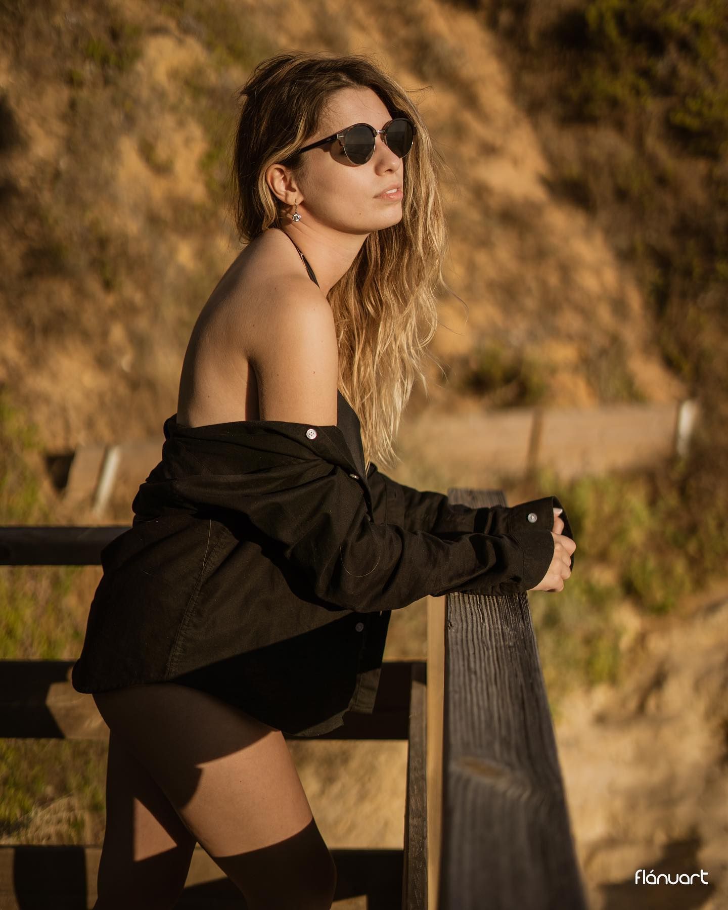 Blonde woman in black off-shoulder shirt and sunglasses posing on wooden fence outdoors.