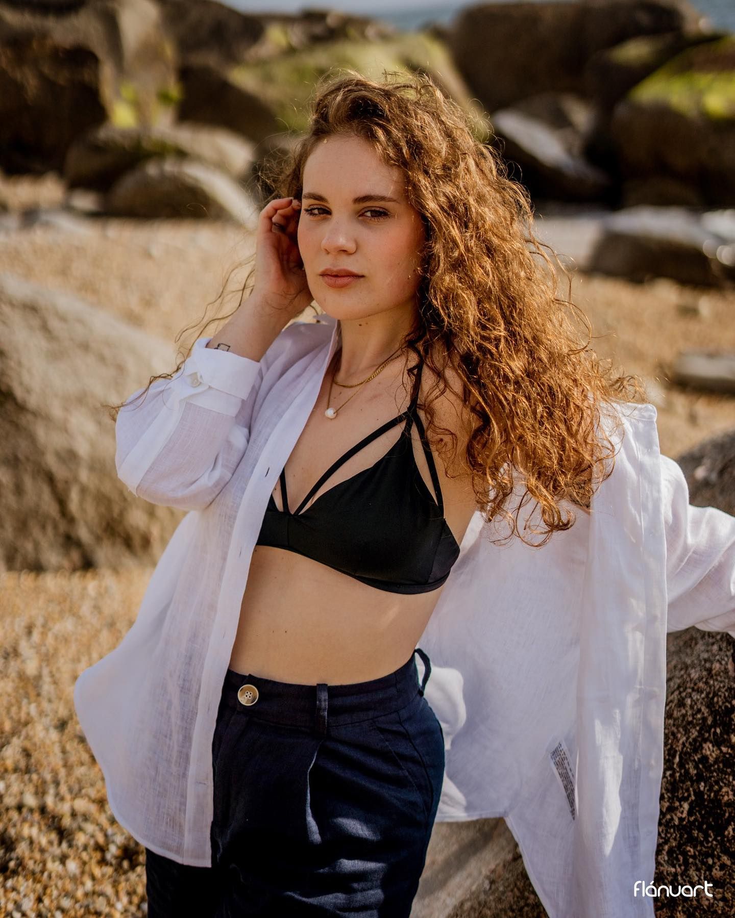 Curly redhead woman in black bralette and open white shirt posing on rocky beach shoreline.