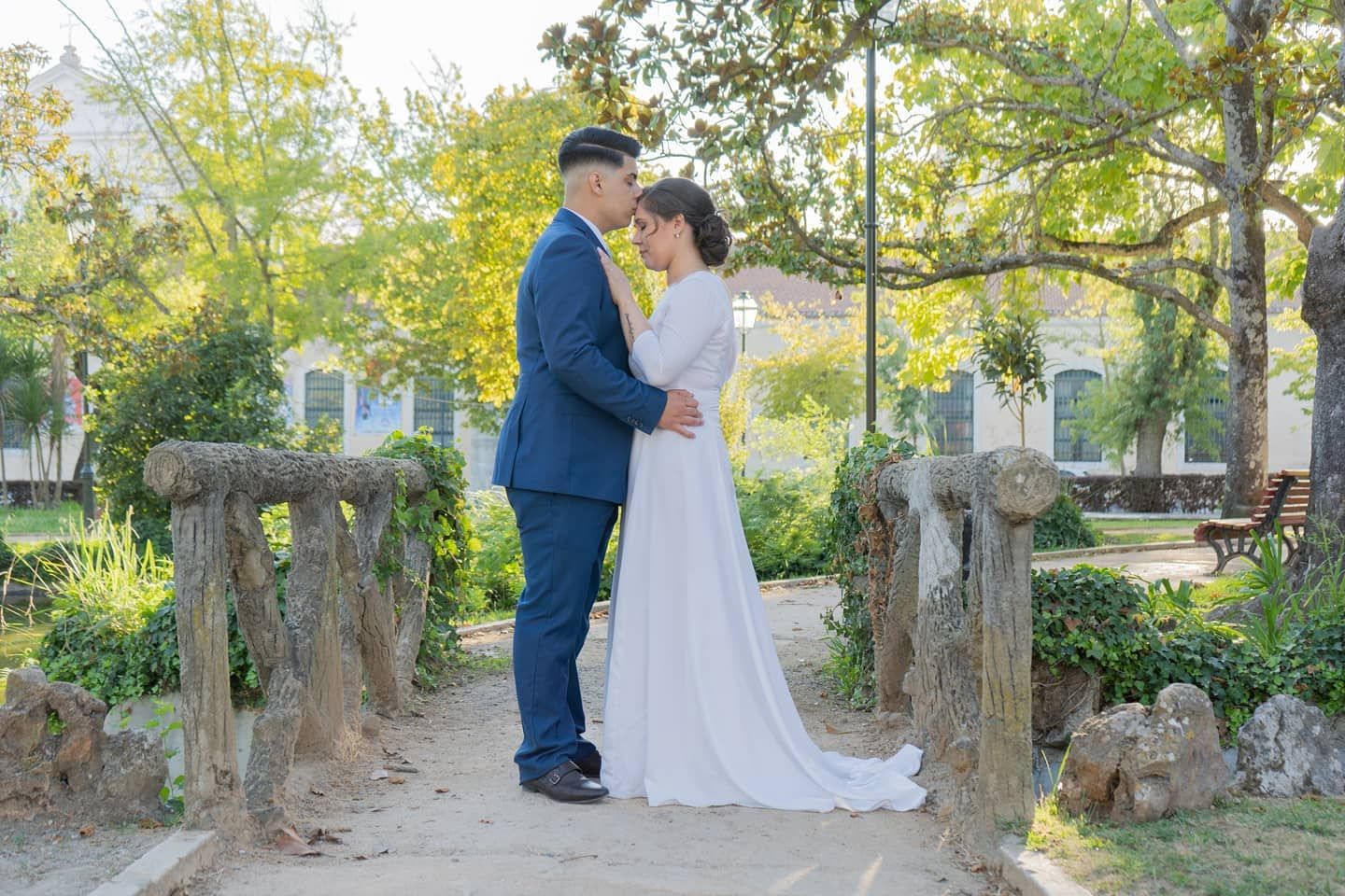 Bride and groom share a romantic moment on a rustic stone bridge surrounded by lush green trees.