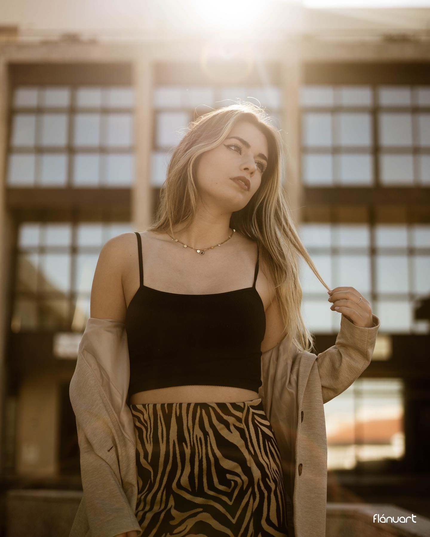 Blonde woman in black crop top, zebra skirt, and beige jacket posing in golden backlight.
