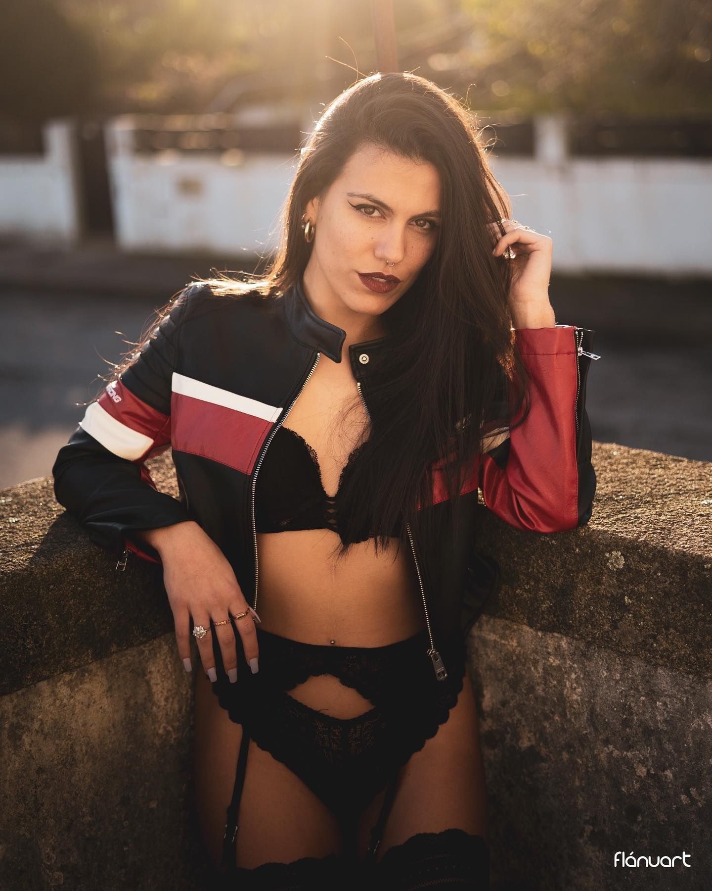 Dark-haired woman in a racing jacket and black lingerie posing outdoors at golden hour.