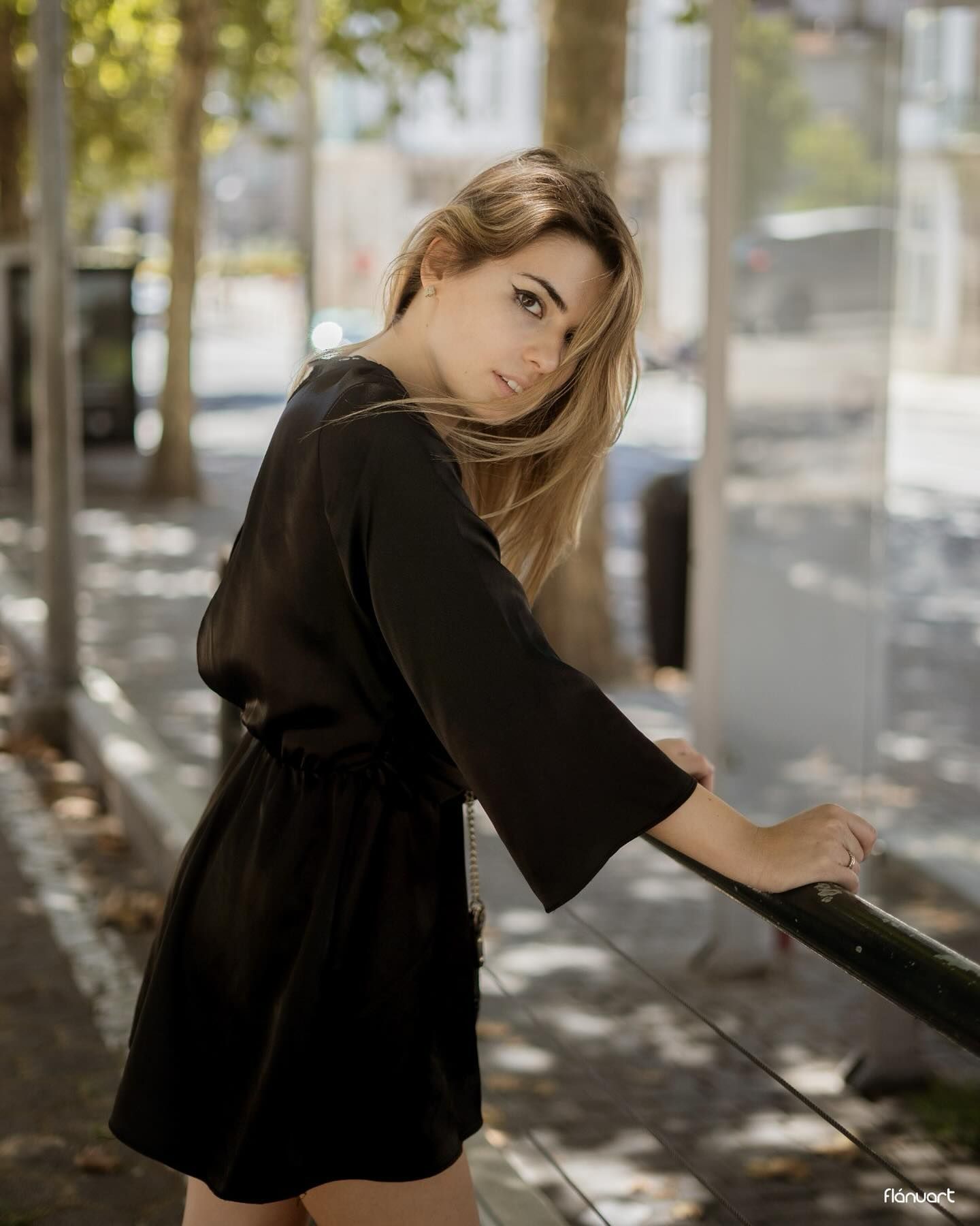 Blonde woman in black mini dress glancing back over shoulder on a sunny city sidewalk.