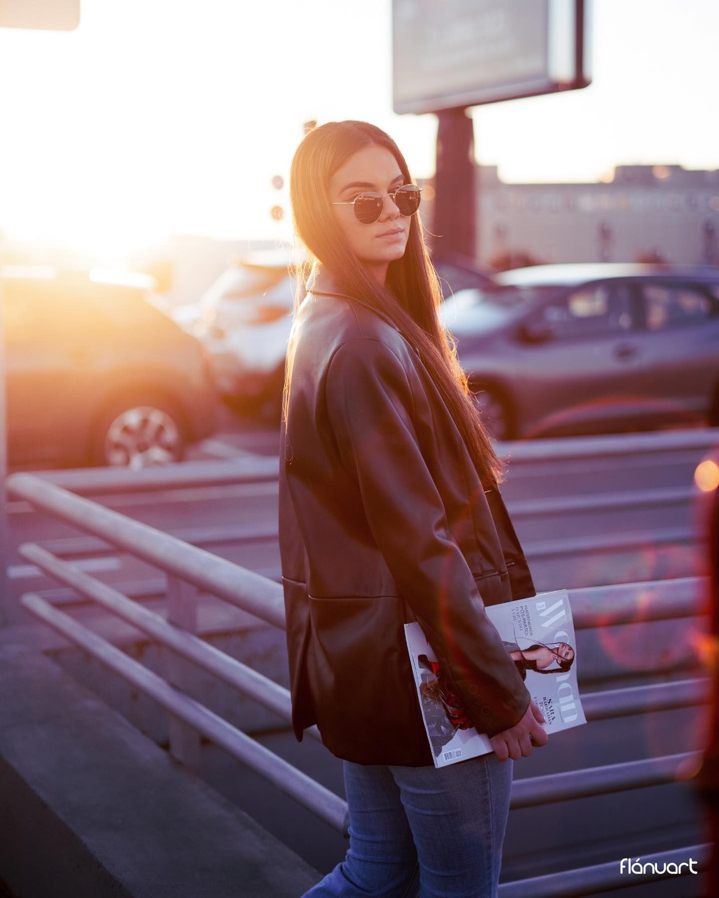 Woman in black leather blazer and jeans holding a magazine, posing at sunset in a parking area.