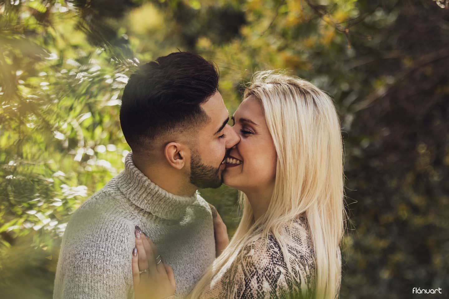 A couple shares a romantic almost-kiss moment surrounded by lush green foliage in a sunny outdoor setting.