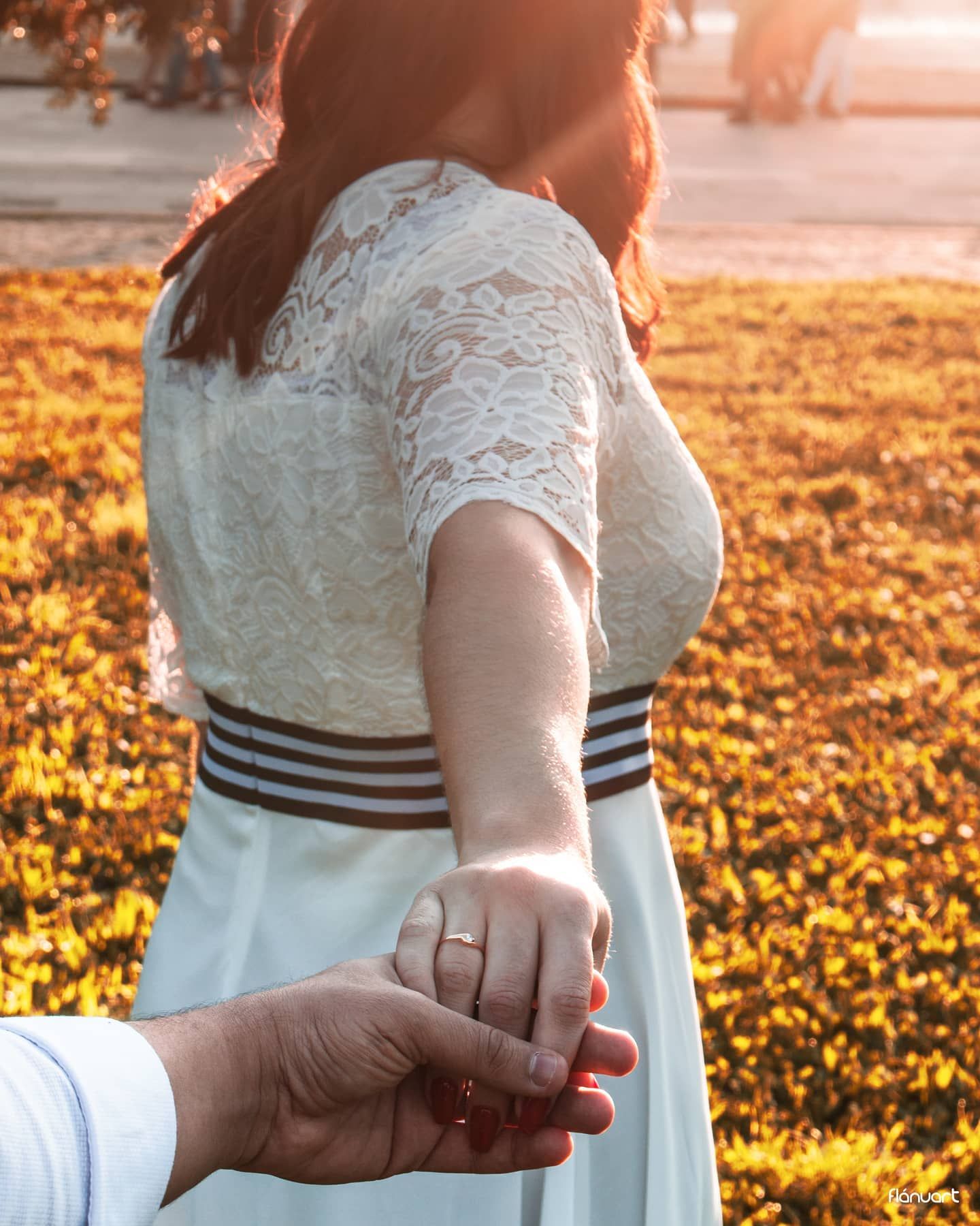 Woman in white lace dress holding hands with partner during golden hour in flower-filled garden.