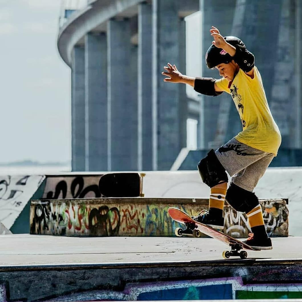 Young skateboarder performing a trick on a ramp at an outdoor skate park near a bridge.