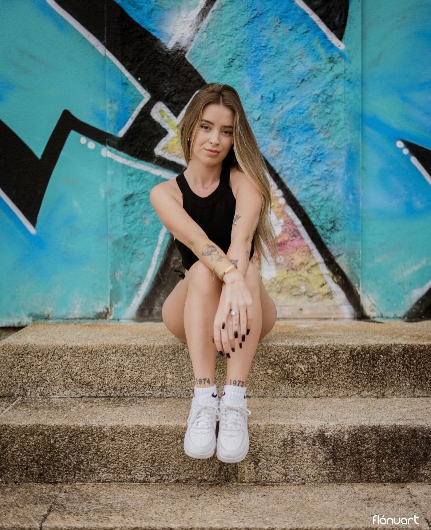 Young woman sitting on graffiti steps in black bodysuit and white sneakers.