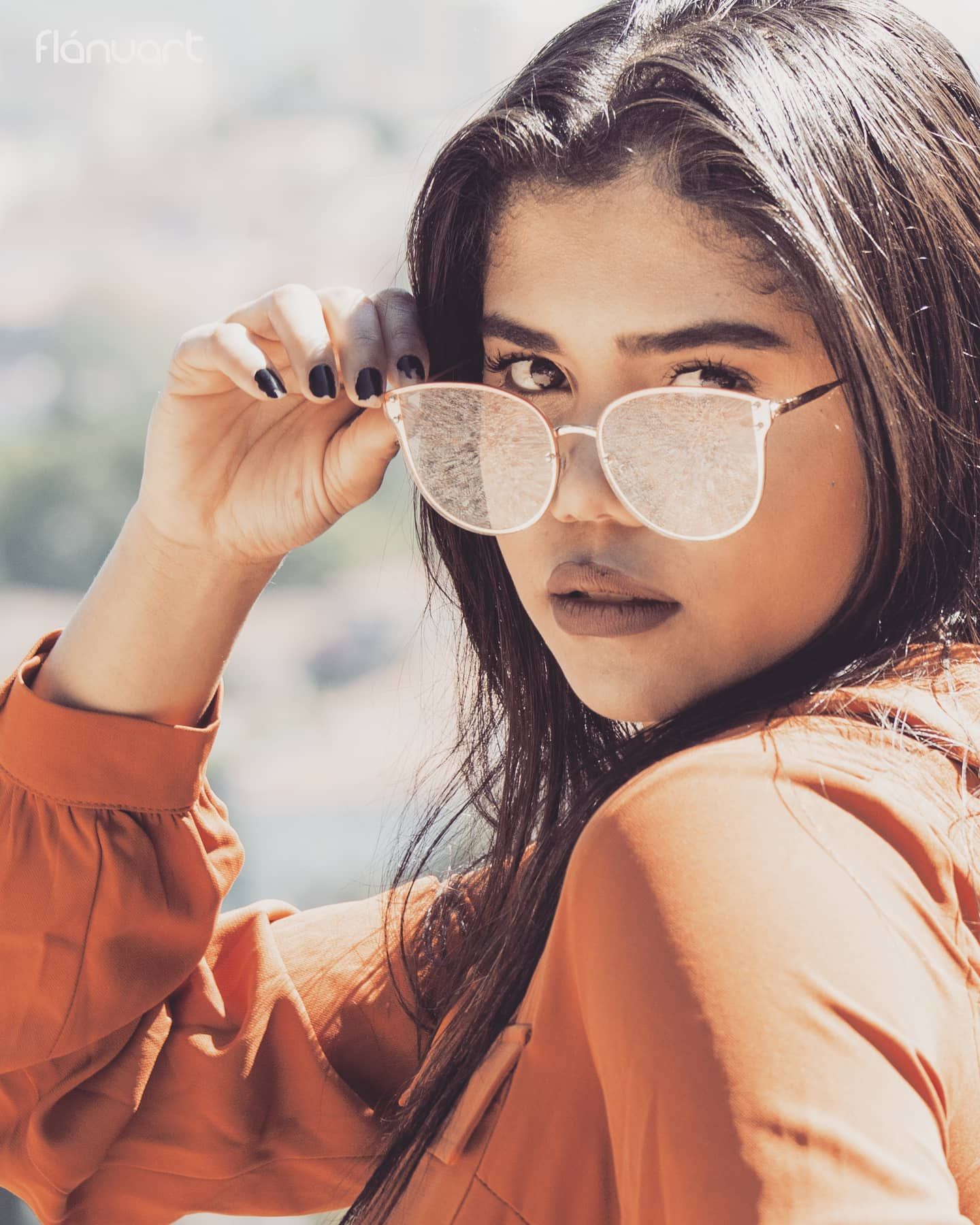 Young woman with dark hair holding white sunglasses wearing an orange top in a close-up portrait.