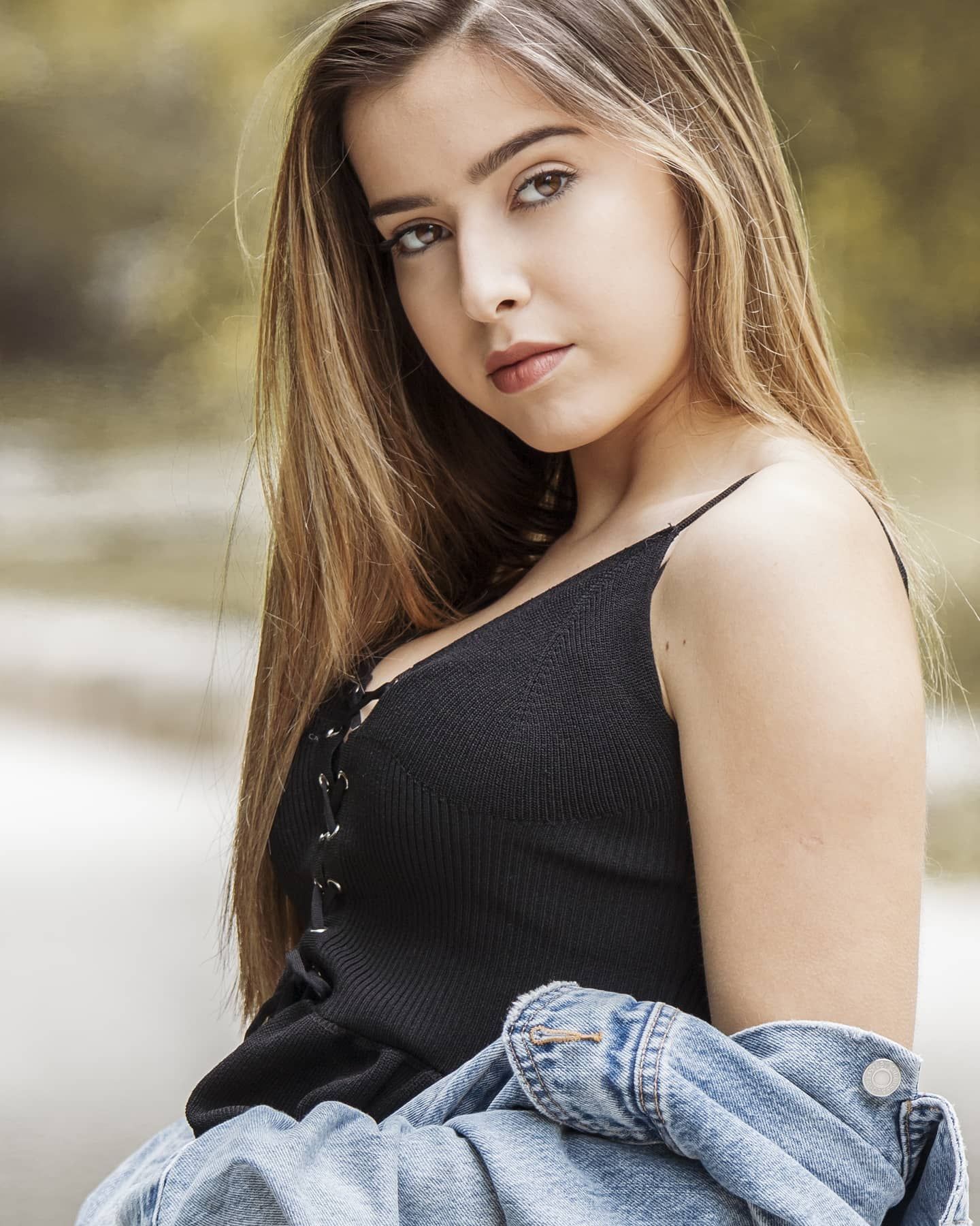 Young woman with long hair in a black lace-up top and denim jacket posing outdoors with soft bokeh background.