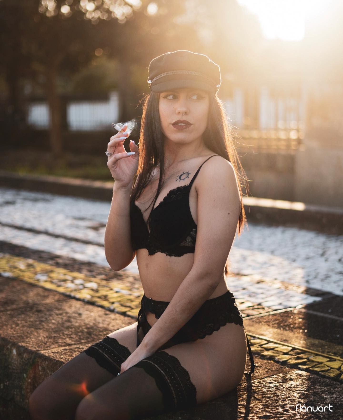 Woman in black lingerie and cap posing outdoors at golden hour with sun flare.
