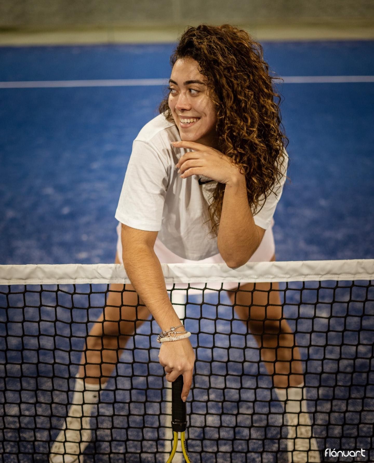 Smiling curly-haired woman leaning on a tennis net holding a racket on a blue court.