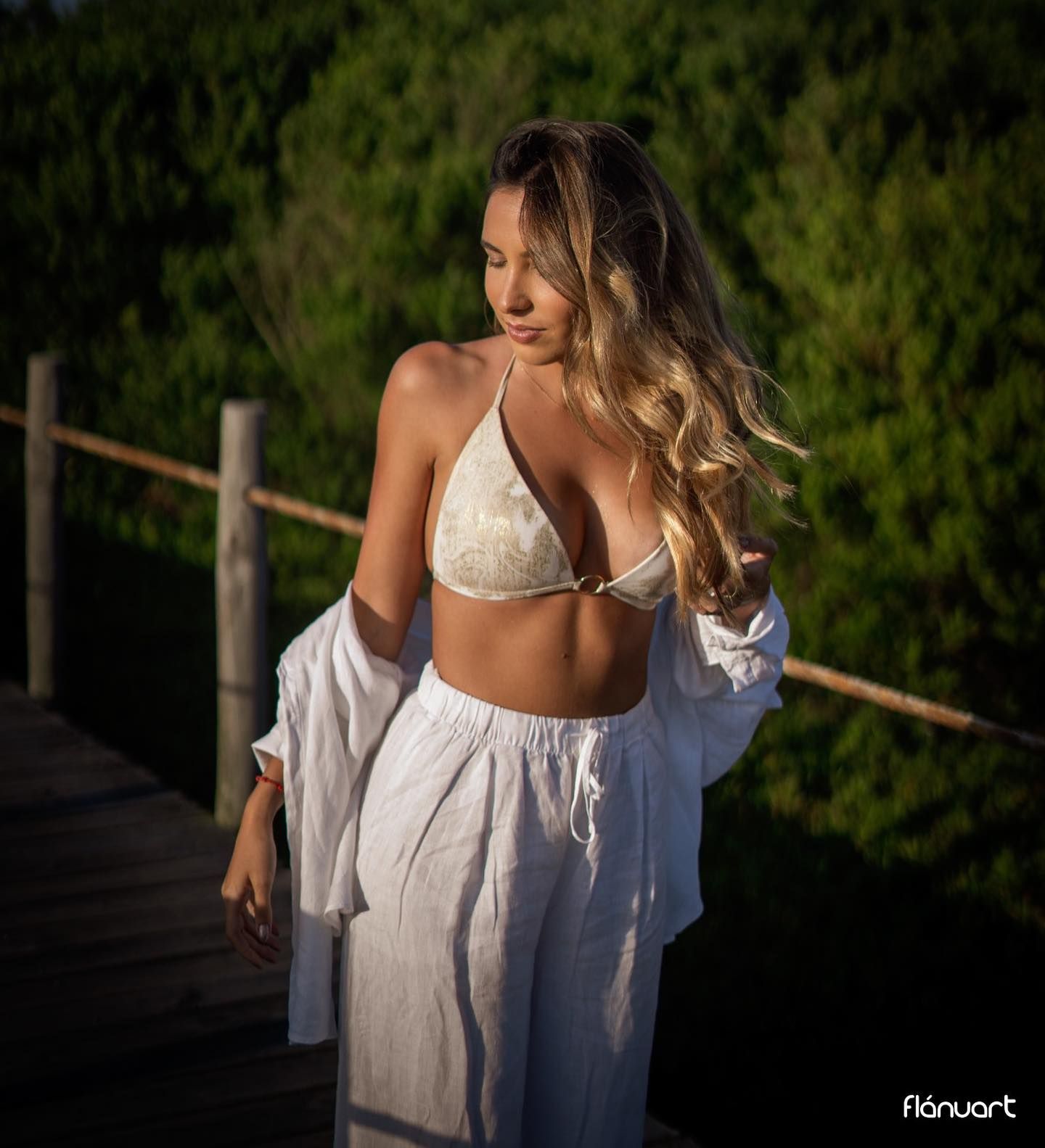 Woman in white bikini top and flowing skirt on wooden bridge surrounded by lush greenery.