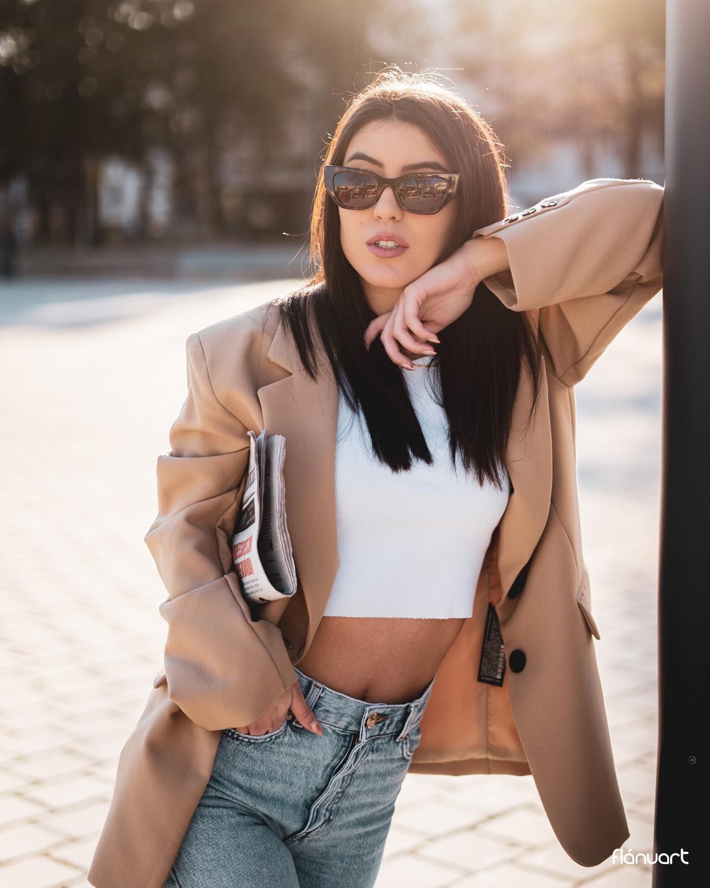 Brunette woman in beige blazer and white crop top posing outdoors in golden sunlight.