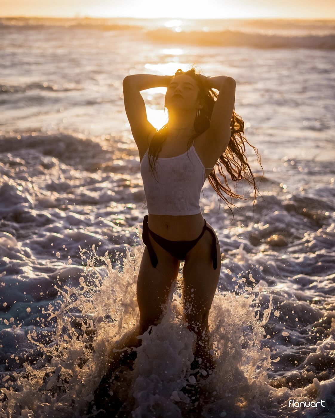 Woman in white crop top and black bikini bottom standing in ocean waves at golden sunset.