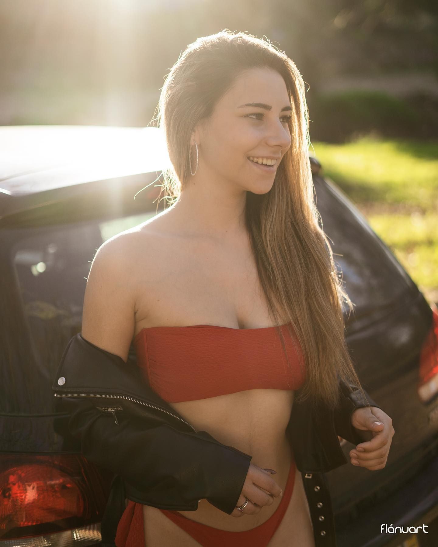 Smiling woman in red bandeau top and leather jacket near car in sunlight.