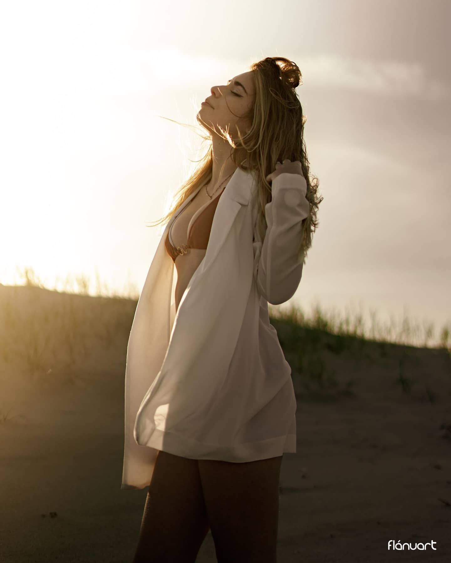 Blonde woman in white oversized shirt posing on sandy beach during golden sunset backlight.