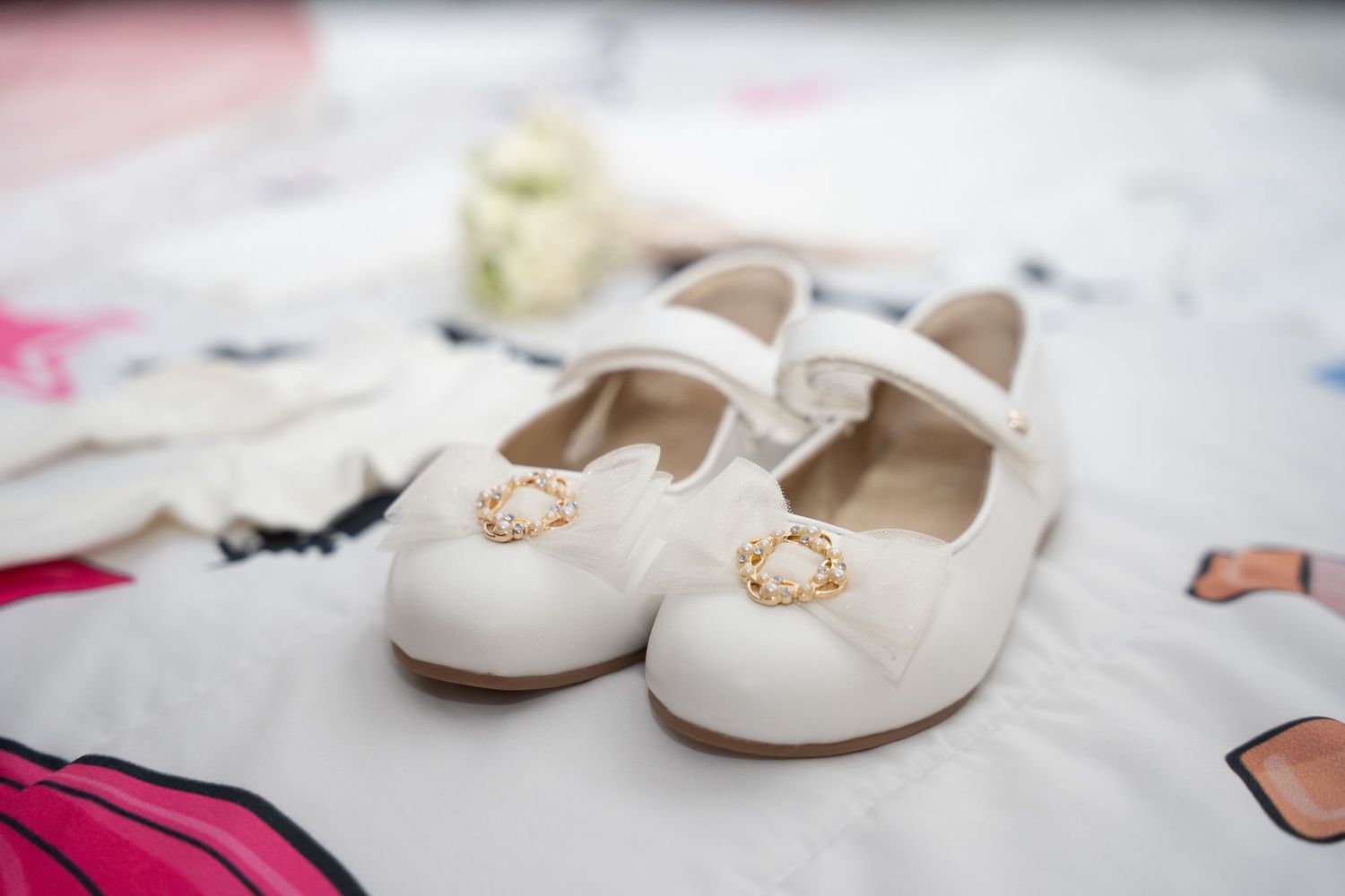 White Mary Jane toddler shoes with gold buckle detail displayed on a floral fabric surface.