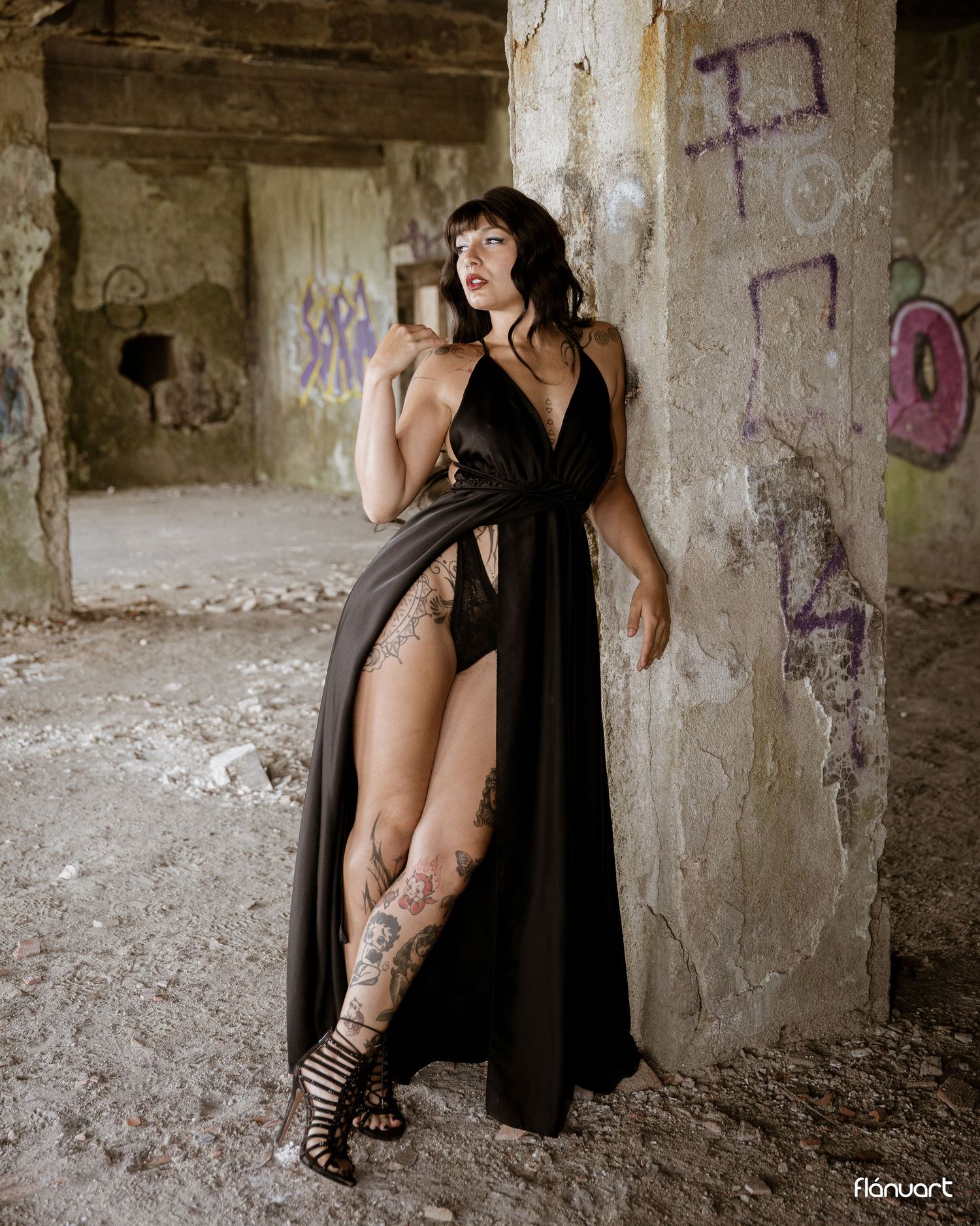 Woman in black slit dress with tattoos poses in abandoned building with graffiti walls.
