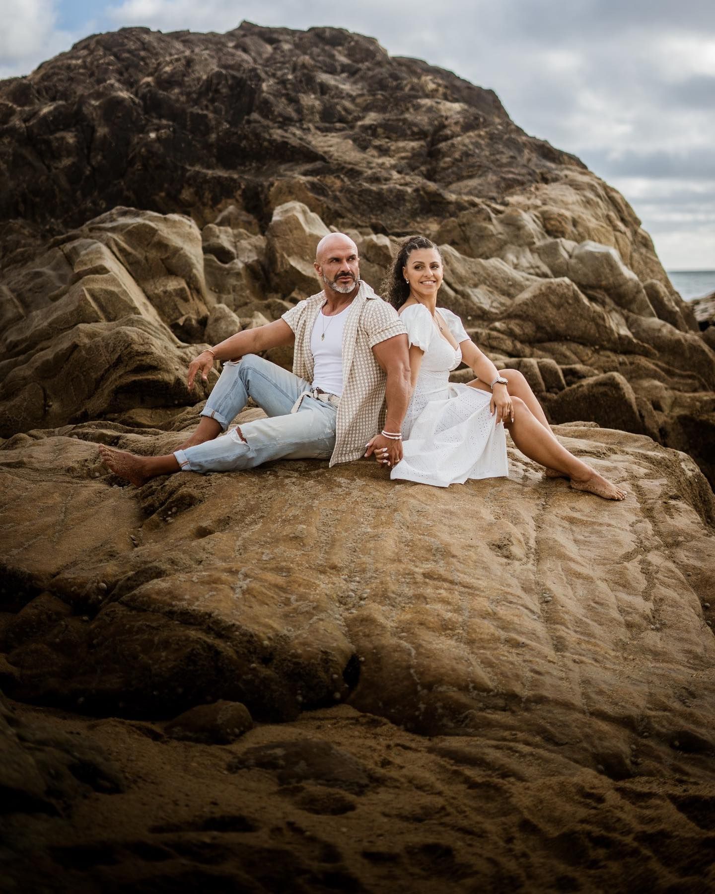 Couple sitting together on large coastal rocks, woman in white dress and man in jeans holding hands.