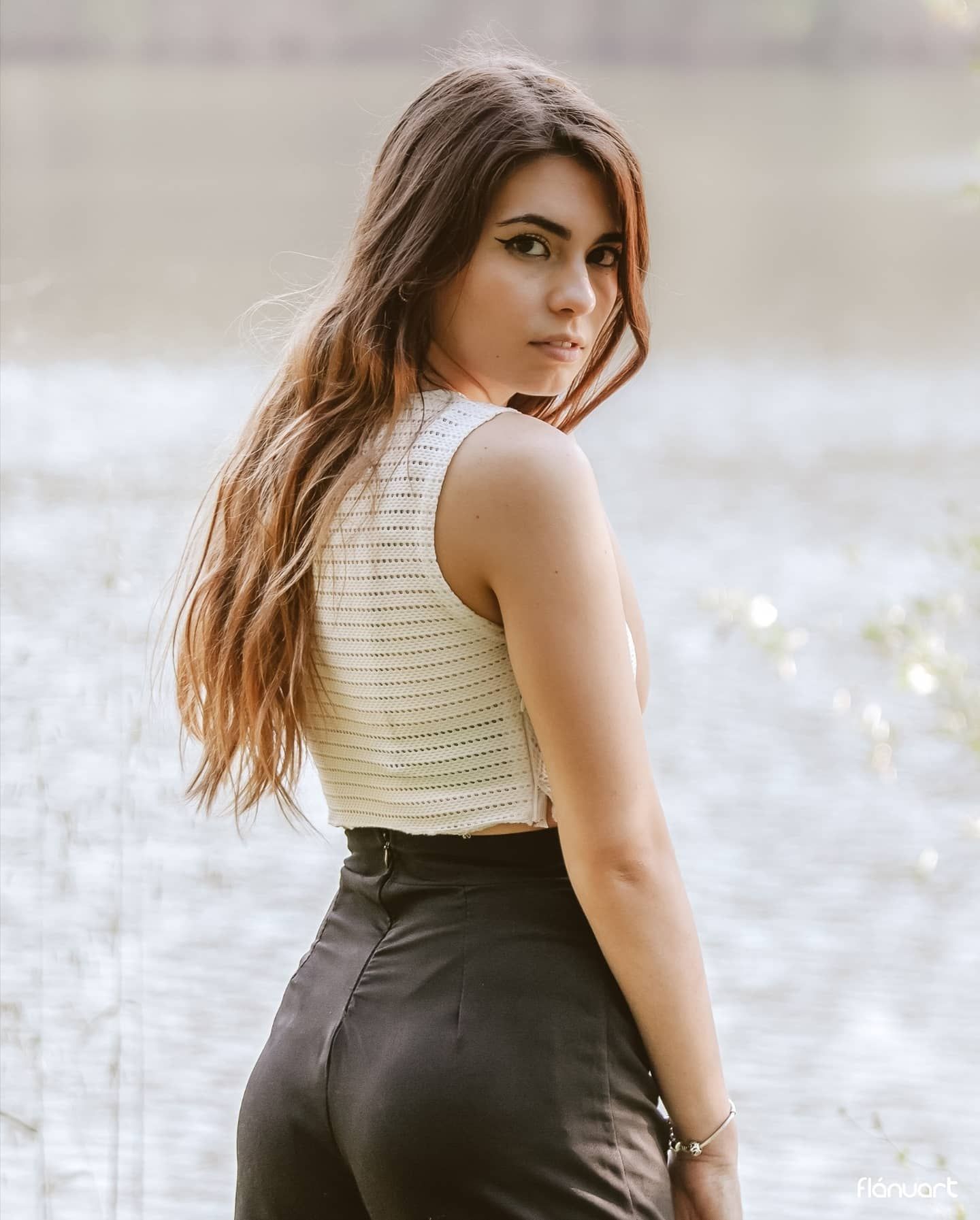 Woman in white knit top and black pants glances back over shoulder near shimmering lake.