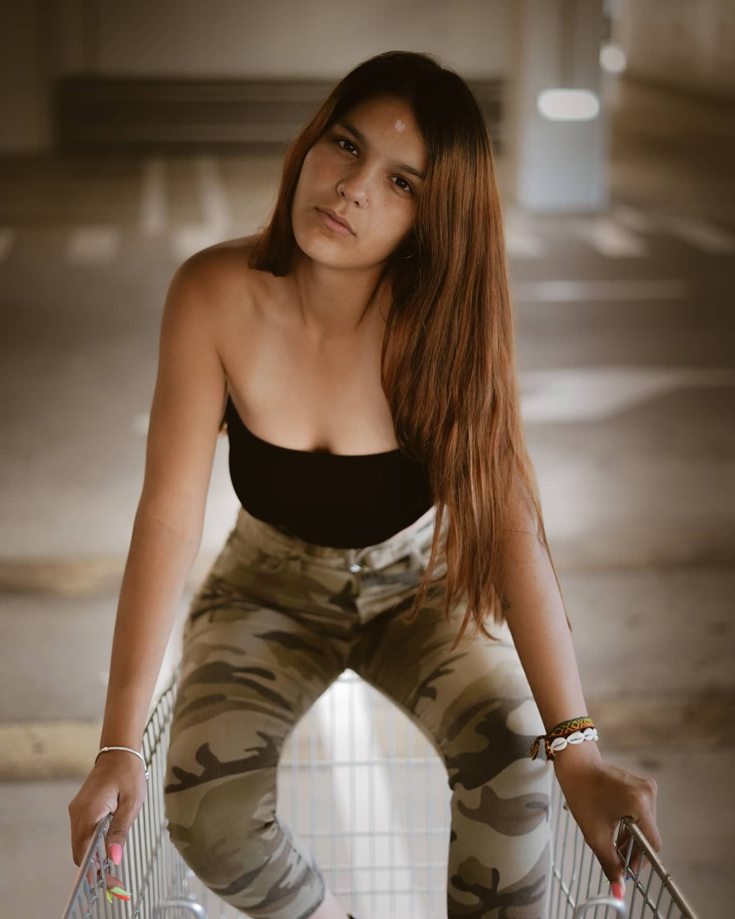Young woman in black tube top and camouflage pants posing with shopping cart in parking garage.