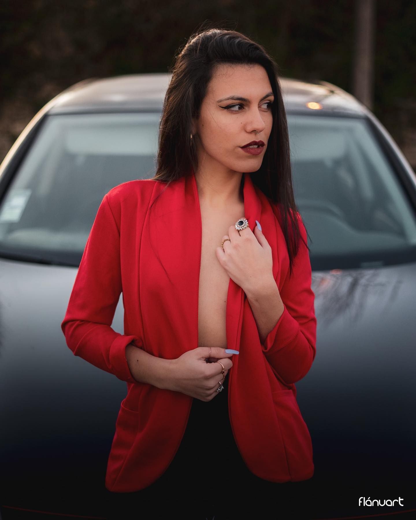 Woman in red blazer posing confidently in front of a dark car outdoors.