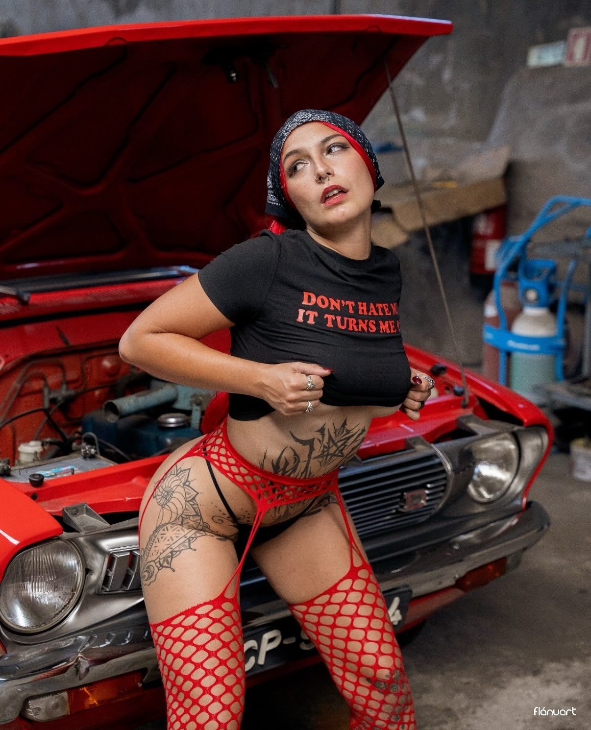 Tattooed woman in crop top and red fishnet stockings poses against a red car in a garage.
