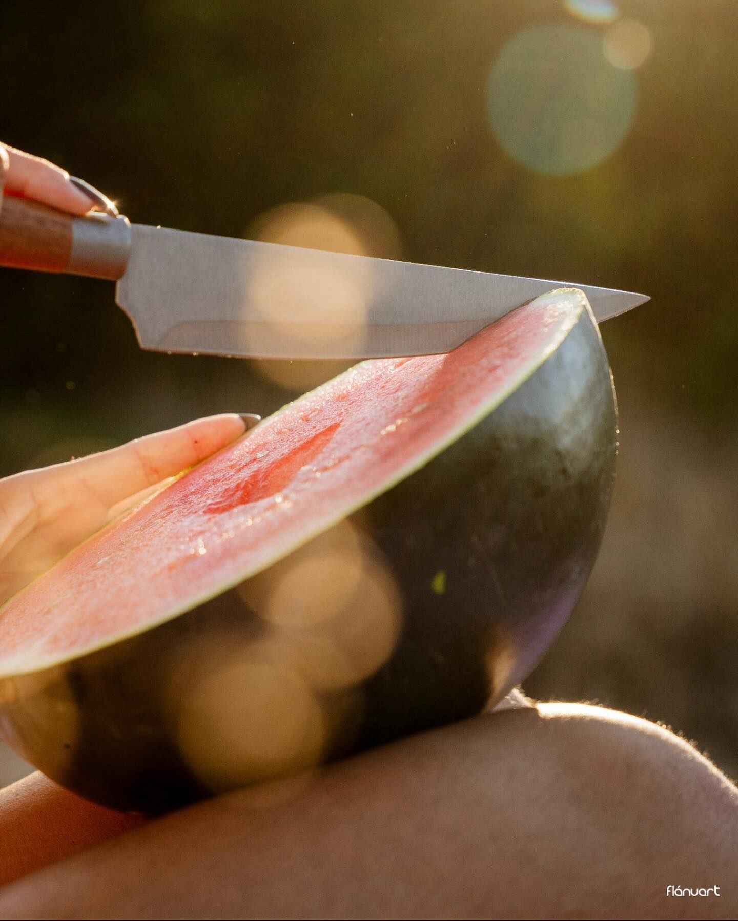 Close-up of hands slicing fresh watermelon with knife in warm golden sunlight outdoors.