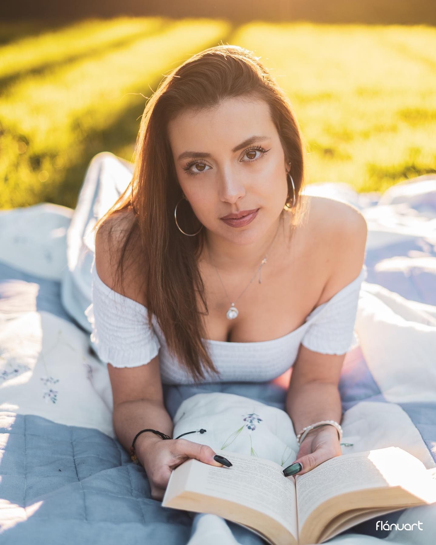 Woman in white off-shoulder dress lying on blanket reading book in golden sunlight.