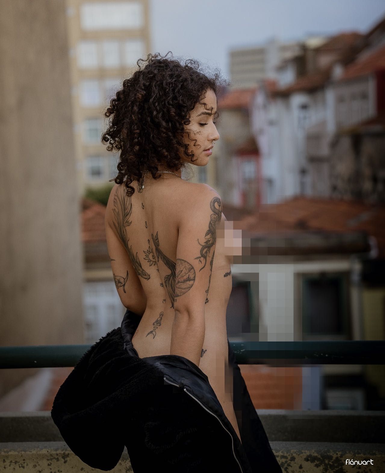 Woman with curly hair and back tattoos posing topless on a balcony with a city skyline behind her.
