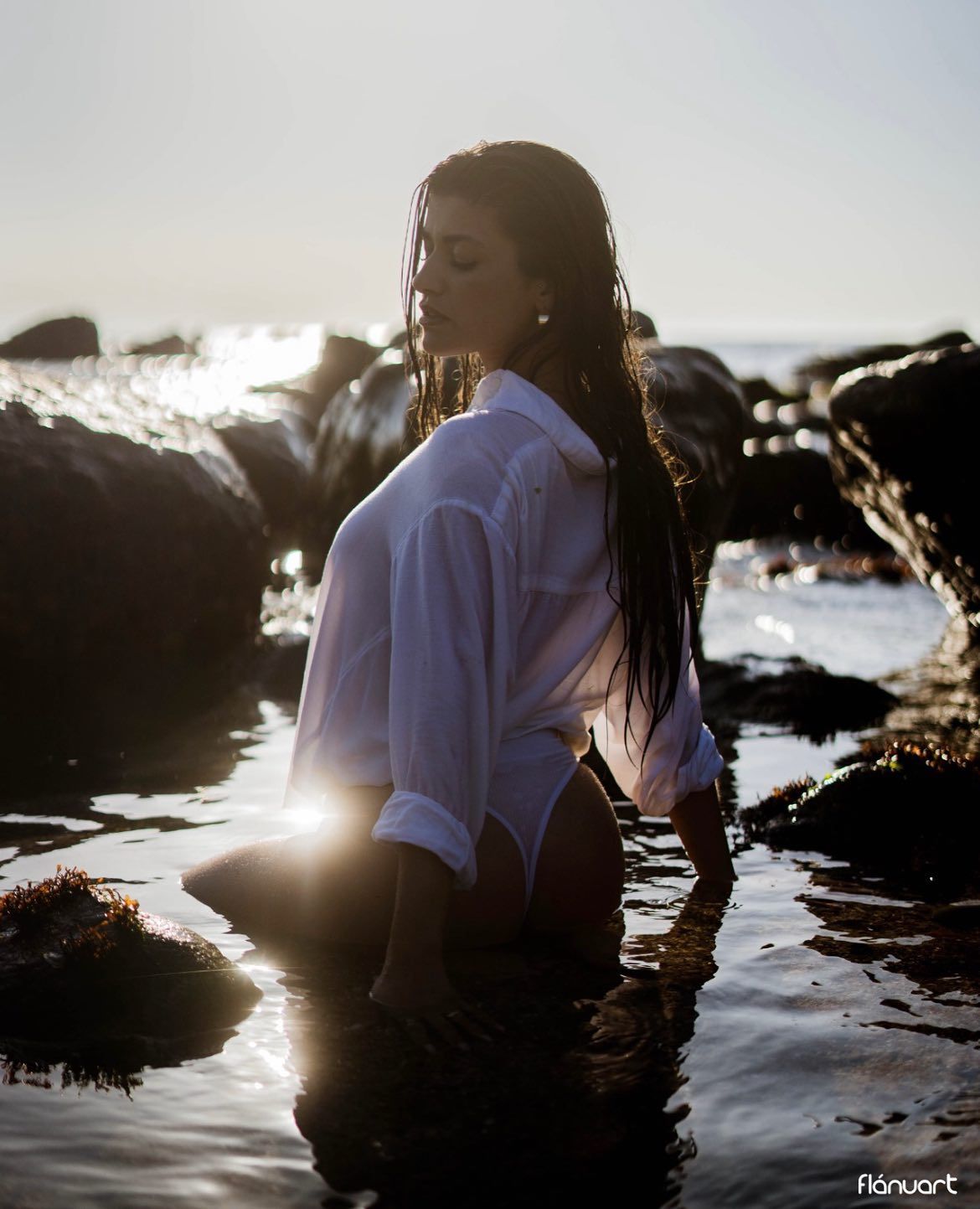 Woman in wet white shirt kneeling in shallow rocky ocean water during golden hour sunset.