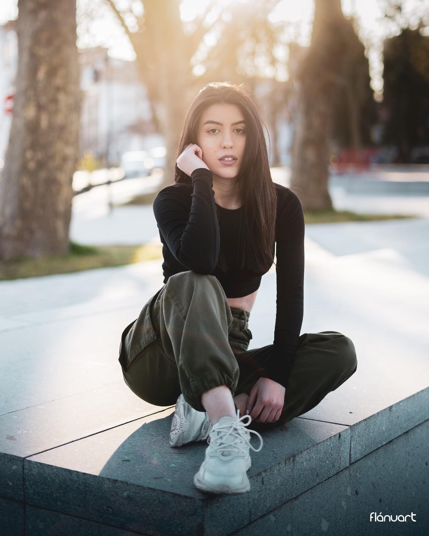 Young woman in black crop top and olive cargo pants sitting cross-legged on a ledge in a sunlit park.