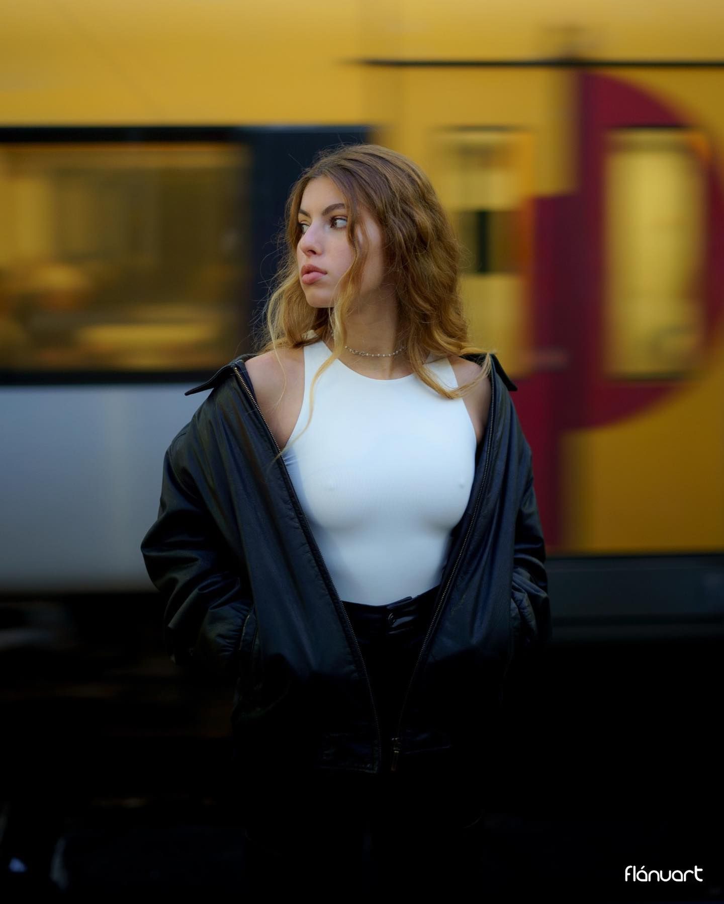 Young woman in white top and black leather jacket poses at a train station with yellow train in background.