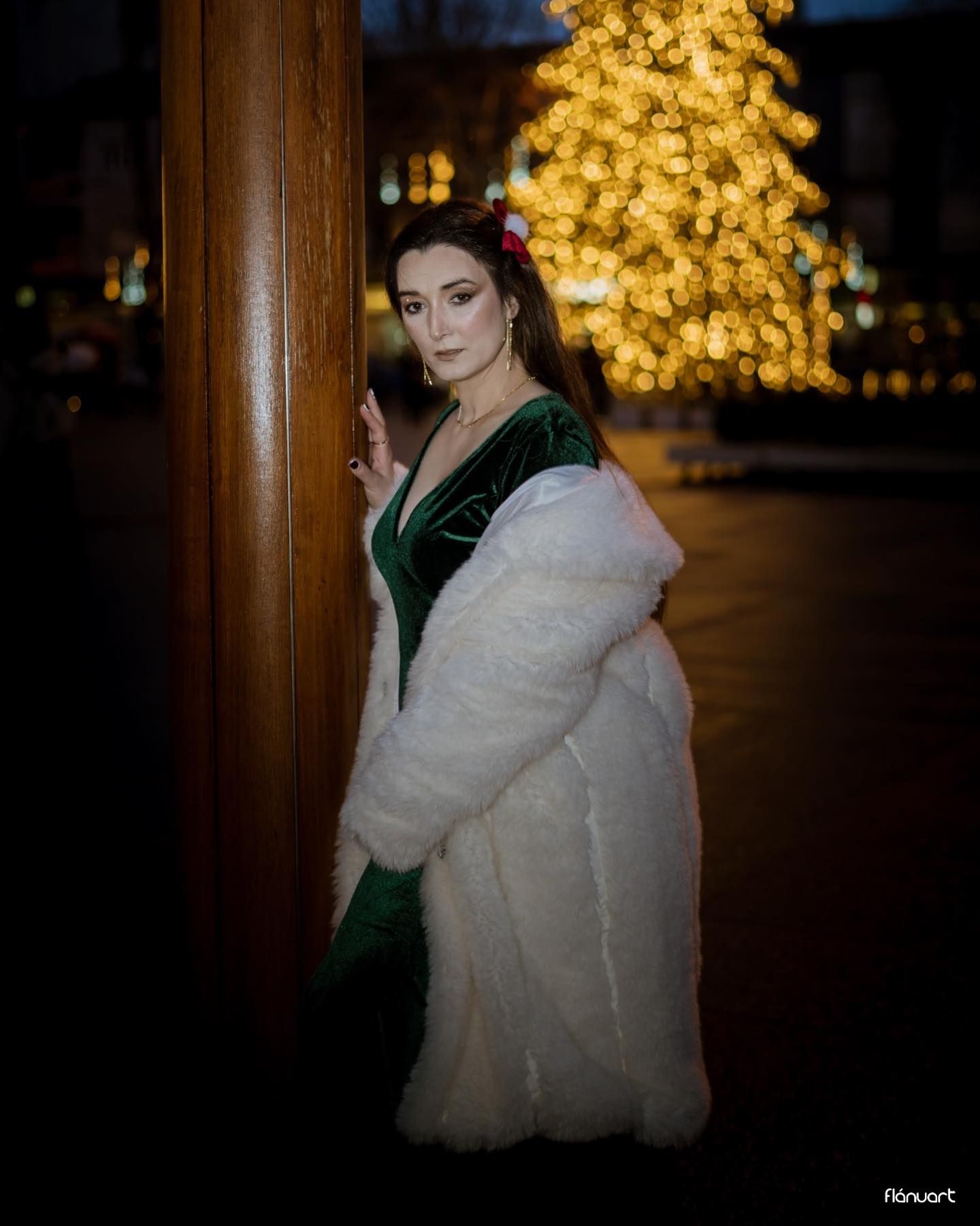 Elegant woman in green velvet dress and white fur coat beside Christmas tree at night.