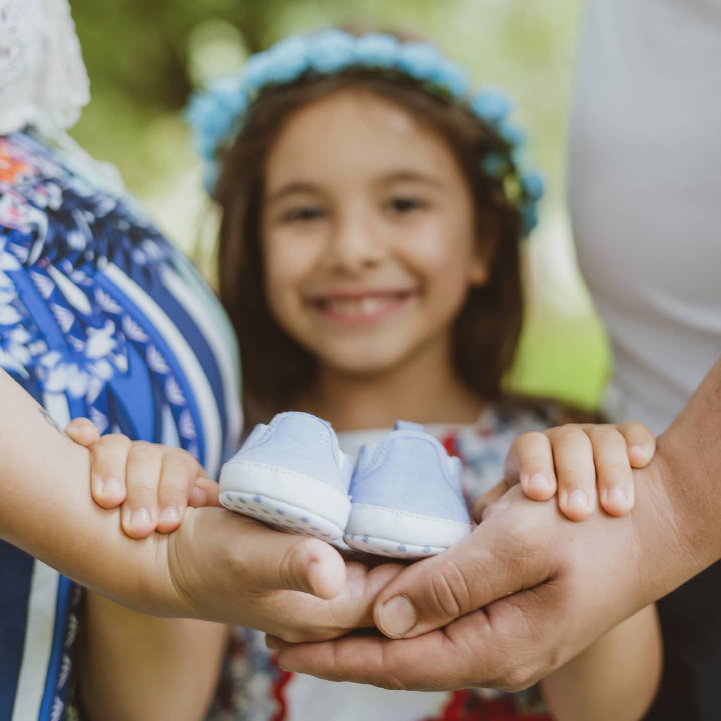 Family hands cradling small blue baby shoes with smiling girl wearing flower crown behind them.