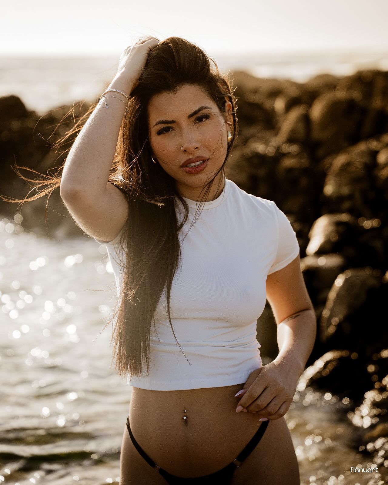 Young woman in white crop top and black bikini bottom posing on rocky beach at sunset.