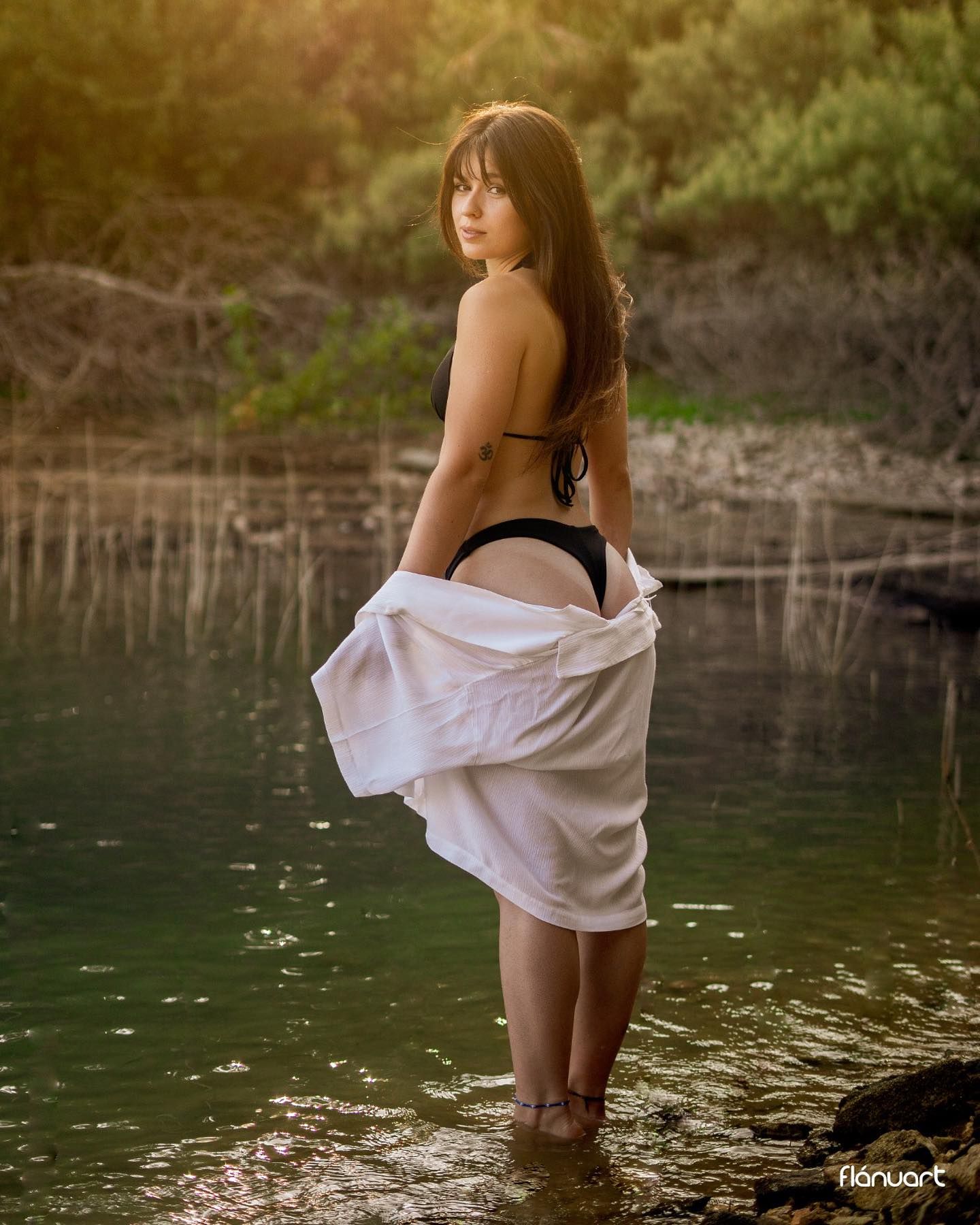 Woman in black bikini and open white shirt posing by sunlit river with lush greenery.
