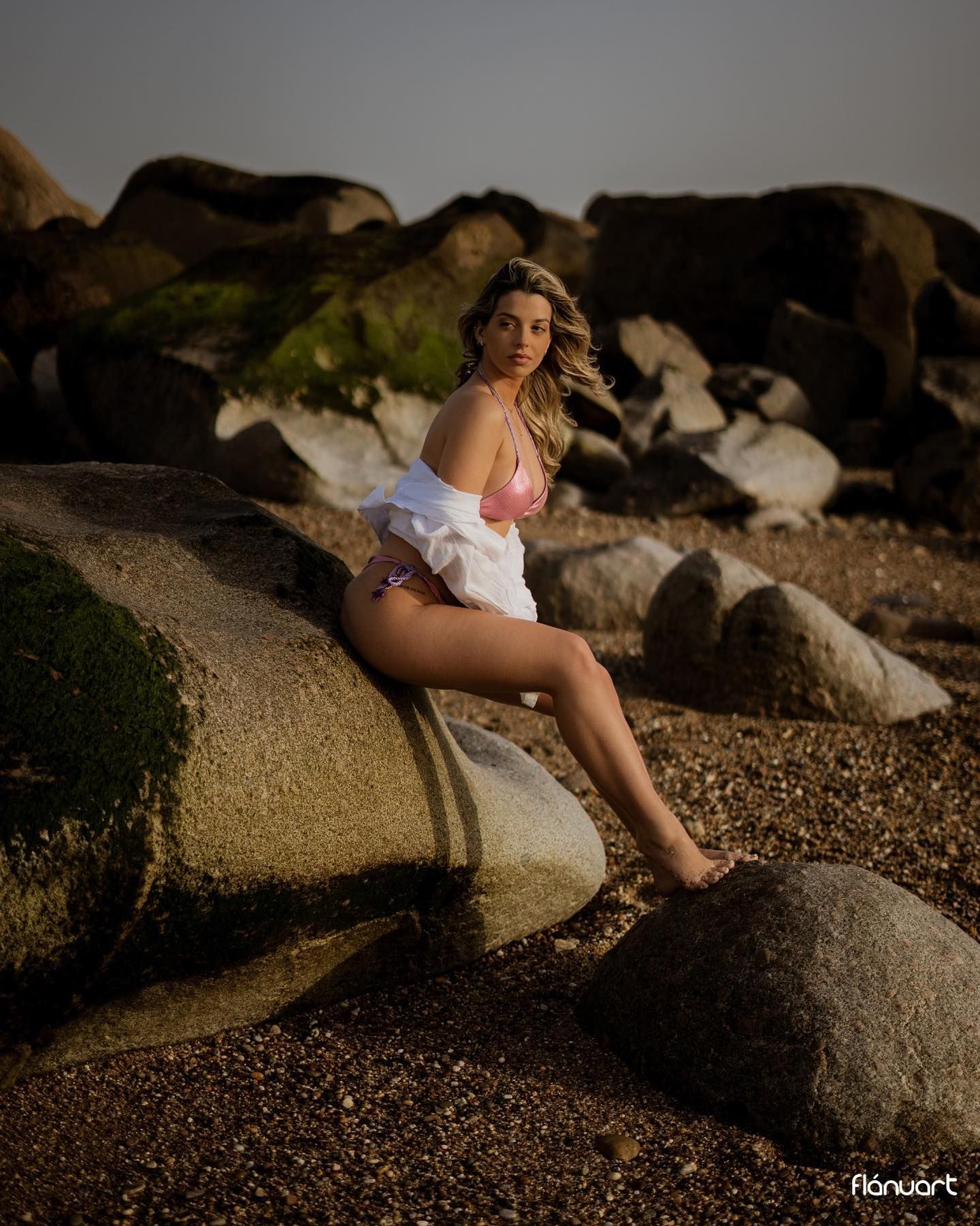 Blonde woman in pink bikini and white shirt posing on rocky beach shore.