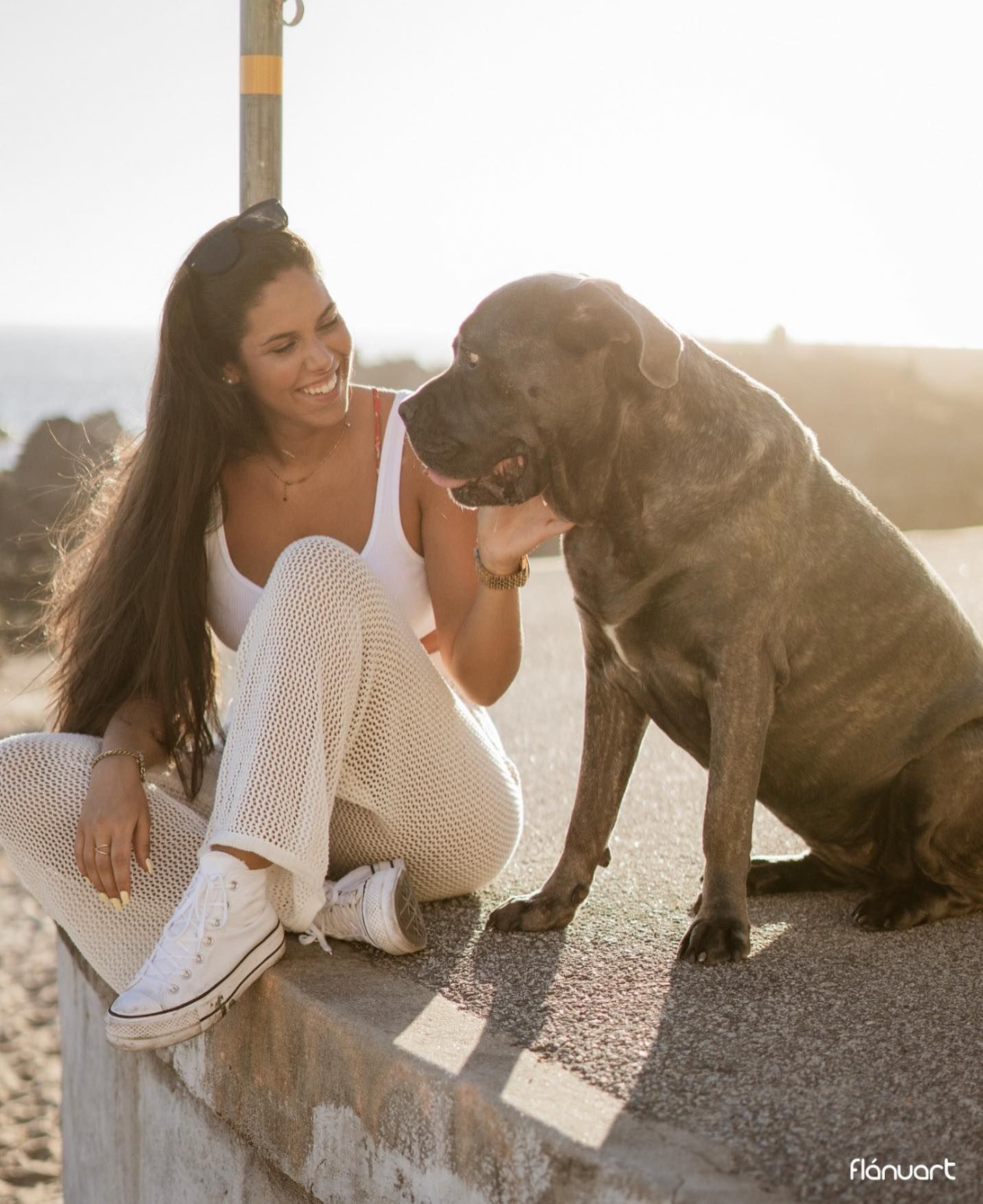 Smiling woman in white outfit sitting with large brindle dog near ocean at sunset.