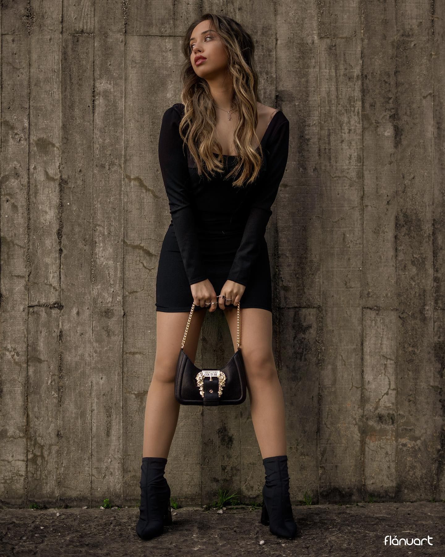 Woman in black off-shoulder mini dress holds designer handbag against concrete wall.