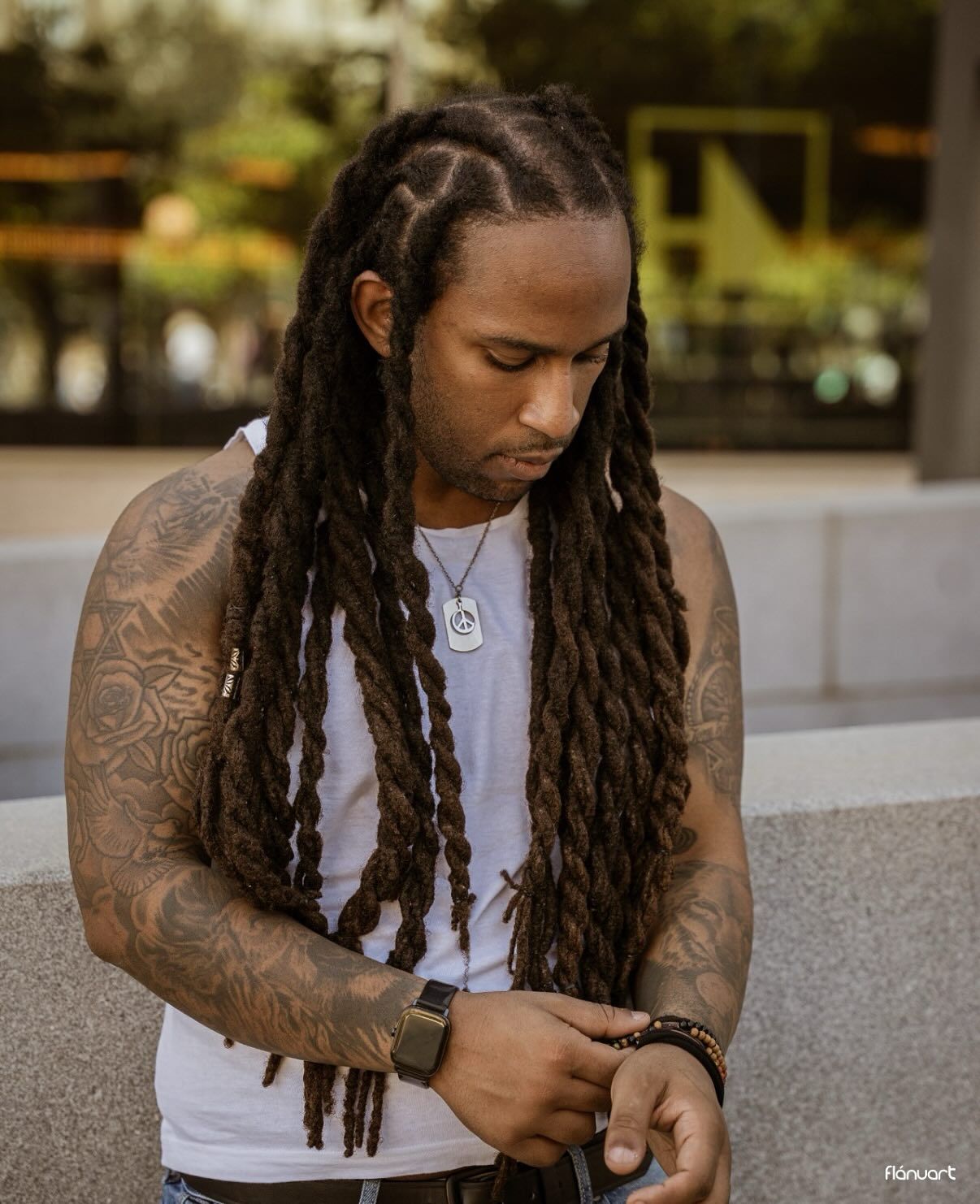 Tattooed man with long dreadlocks wearing a white tank top and beaded bracelet looking down outdoors.