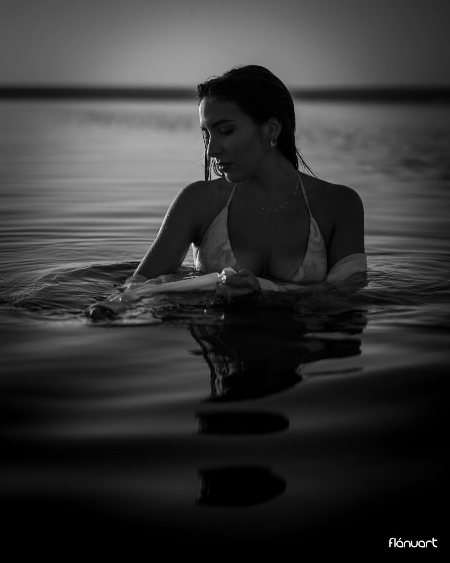 Black and white photo of woman in bikini standing in calm water at dusk.