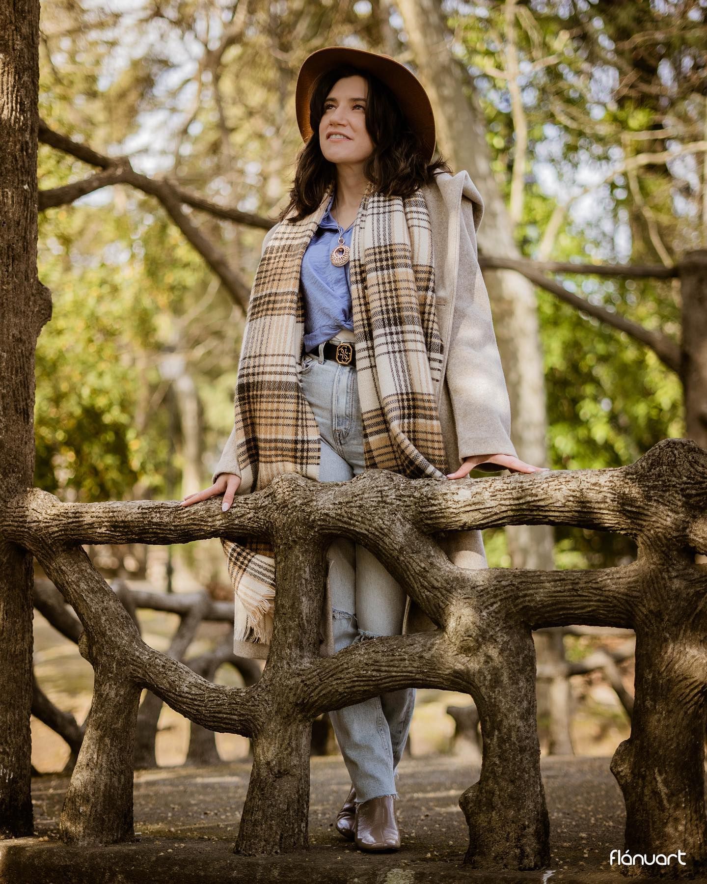 Smiling woman in plaid poncho and hat standing on tree branches in sunny forest.