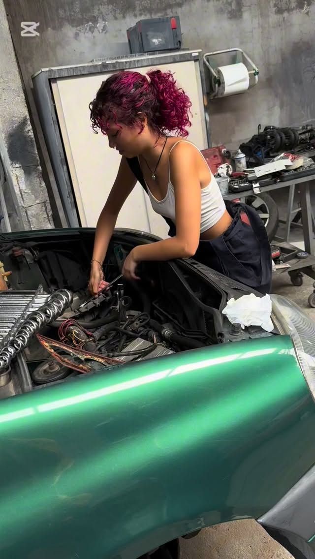 Woman with pink curly hair works on a car engine in a mechanic garage wearing casual clothing.